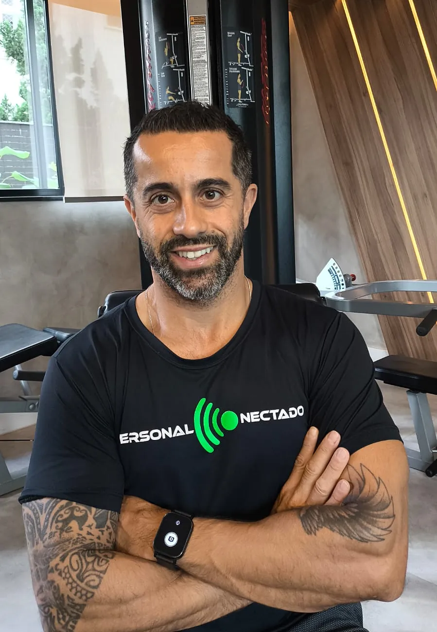 Gustavo sorrindo, de braços cruzados, em frente a um equipamento de musculação